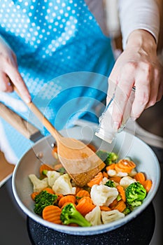 Young housewife holding wooden spoon and adding salt over various vegetables meal in pan. Vegetarian food cooking