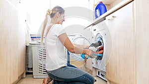 Young housewife sitting on floor and pulling heap of clean clothes out of washing machine