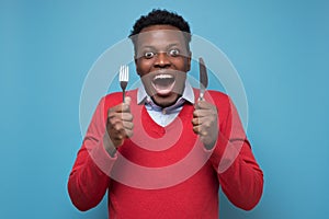 Young hungry african man holding a fork and a knife waiting for delicious meal.