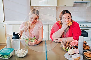 Young hungry slim and overweight women in kitchen eating food. Healthy and unhealthy meal. Salad vs burger. Body