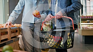 Young husband holding basket full of various items while wife putting in oranges