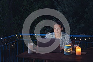 Young hysster woman working on a laptop in the evening on the open terrace of her country house, cozy with candles and lanterns