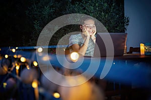 Young hysster woman working on a laptop in the evening on the open terrace of her country house, cozy with candles and lanterns