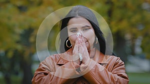 Young ill hispanic woman standing outdoors sneezing into handkerchief wiping nose feeling unwell illness seasonal cold