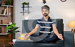 Young indian boy meditating with eyes closed before reading while sitting on sofa at home - concept of stress relief