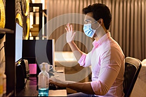 Young Indian businessman with mask video calling while working overtime at home during quarantine