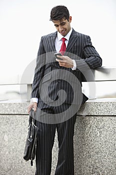 Young Indian businessman texting through cell phone while holding laptop bag at parapet