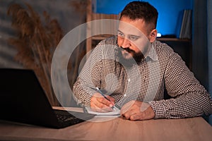 Young indian businessman using a laptop and writing notes while working overtime at his home office desk late in the