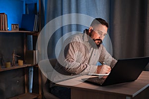 Young indian businessman using a laptop and writing notes while working overtime at his office desk late in the evening