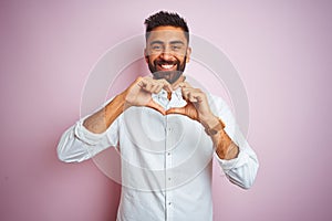 Young indian businessman wearing elegant shirt standing over isolated pink background smiling in love doing heart symbol shape