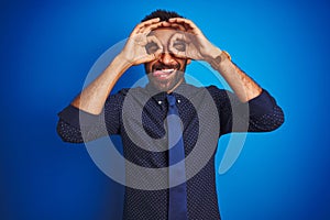 Young indian businessman wearing elegant shirt and tie standing over isolated blue background doing ok gesture like binoculars
