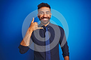 Young indian businessman wearing elegant shirt and tie standing over isolated blue background smiling doing phone gesture with