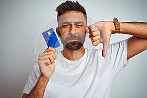 Young indian customer man holding credit card standing over isolated white background with angry face, negative sign showing