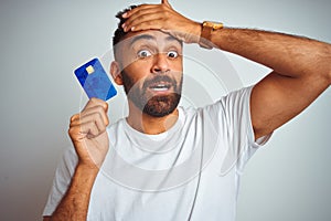 Young indian customer man holding credit card standing over isolated white background stressed with hand on head, shocked with