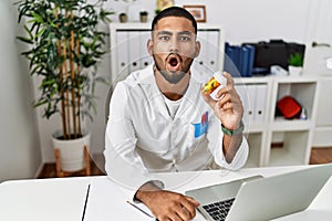 Young indian doctor holding prescription pills scared and amazed with open mouth for surprise, disbelief face