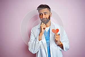 Young indian doctor man holding paper heart standing over isolated pink background serious face thinking about question, very