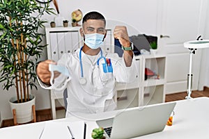 Young indian doctor offering safety mask annoyed and frustrated shouting with anger, yelling crazy with anger and hand raised