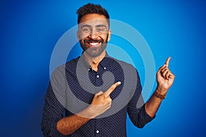 Young indian elegant man wearing shirt standing over isolated blue background smiling and looking at the camera pointing with two