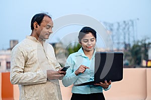 Young Indian female in formal dress with laptop showing something to a middle aged man on internet, The man using his smartphone