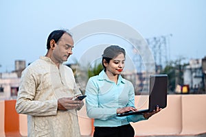 Young Indian female in formal dress with laptop showing something to a middle aged man on internet, The man using his smartphone