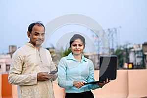 Young Indian female in formal dress with laptop showing something to a middle aged man on internet, The man using his smartphone