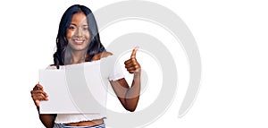 Young indian girl holding blank empty banner smiling happy and positive, thumb up doing excellent and approval sign