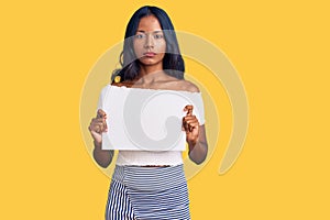 Young indian girl holding blank empty banner thinking attitude and sober expression looking self confident