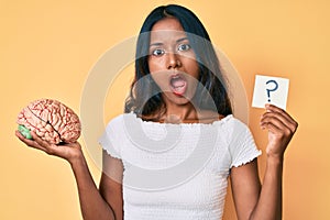 Young indian girl holding brain and question mark reminder in shock face, looking skeptical and sarcastic, surprised with open