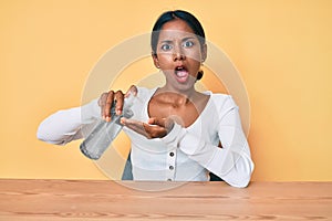 Young indian girl sitting on the table cleaning hand using sanitizer gel in shock face, looking skeptical and sarcastic, surprised