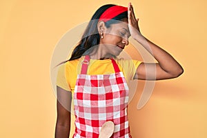 Young indian girl wearing baker apron surprised with hand on head for mistake, remember error