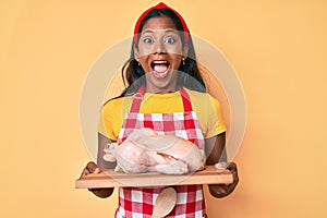 Young indian girl wearing cook apron and holding chicken celebrating crazy and amazed for success with open eyes screaming excited