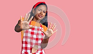 Young indian girl wearing professional baker apron afraid and terrified with fear expression stop gesture with hands, shouting in