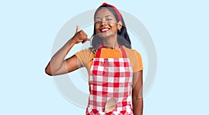 Young indian girl wearing professional baker apron smiling doing phone gesture with hand and fingers like talking on the telephone