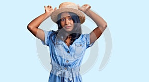 Young indian girl wearing summer hat doing bunny ears gesture with hands palms looking cynical and skeptical