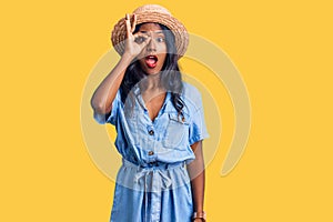 Young indian girl wearing summer hat doing ok gesture shocked with surprised face, eye looking through fingers