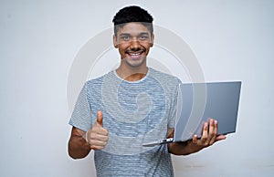 `Young indian handsome boy showing thumbs up and smiling while looking into the camera, holding laptop in his other hand