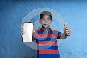 Young indian kid holding a phone with white screen and showing thumbs up in the camera, standing against a blue background