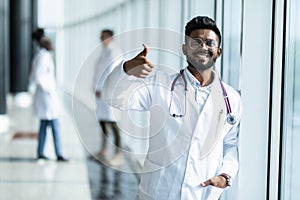 Young indian male doctor with thumbs up in white uniform with collegues on the background