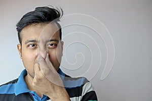 A young indian male showing the gesture of foul smell pinching nose with hands and frowning isolated on grey with copy