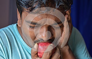 Young Indian man bits an apple and feels pain with sensitive teeth on color background