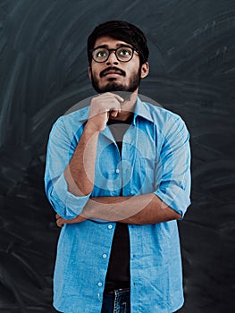A young Indian man in a blue shirt and glasses poses thoughtfully in front of school board