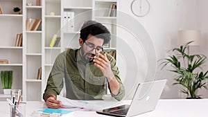 A young Indian man businessman sits at the table, argues on video call on laptop with colleagues, shows graphs