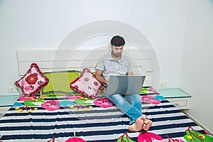 Young indian man busy working on his laptop doing office work while relaxing on bed in bedroom, freelancer working from home.