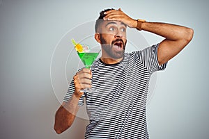 Young indian man drinking cocktail with alcohol standing over isolated white background stressed with hand on head, shocked with