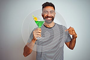 Young indian man drinking cocktail with alcohol standing over isolated white background with surprise face pointing finger to
