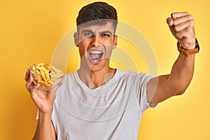 Young indian man holding bowl with dry pasta standing over isolated yellow background annoyed and frustrated shouting with anger,
