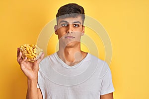 Young indian man holding bowl with dry pasta standing over isolated yellow background with a confident expression on smart face