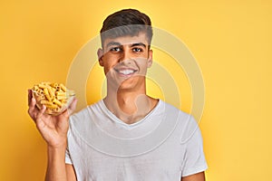 Young indian man holding bowl with dry pasta standing over isolated yellow background with a happy face standing and smiling with