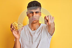 Young indian man holding bowl with dry pasta standing over isolated yellow background pointing with finger to the camera and to