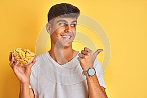 Young indian man holding bowl with dry pasta standing over isolated yellow background pointing and showing with thumb up to the